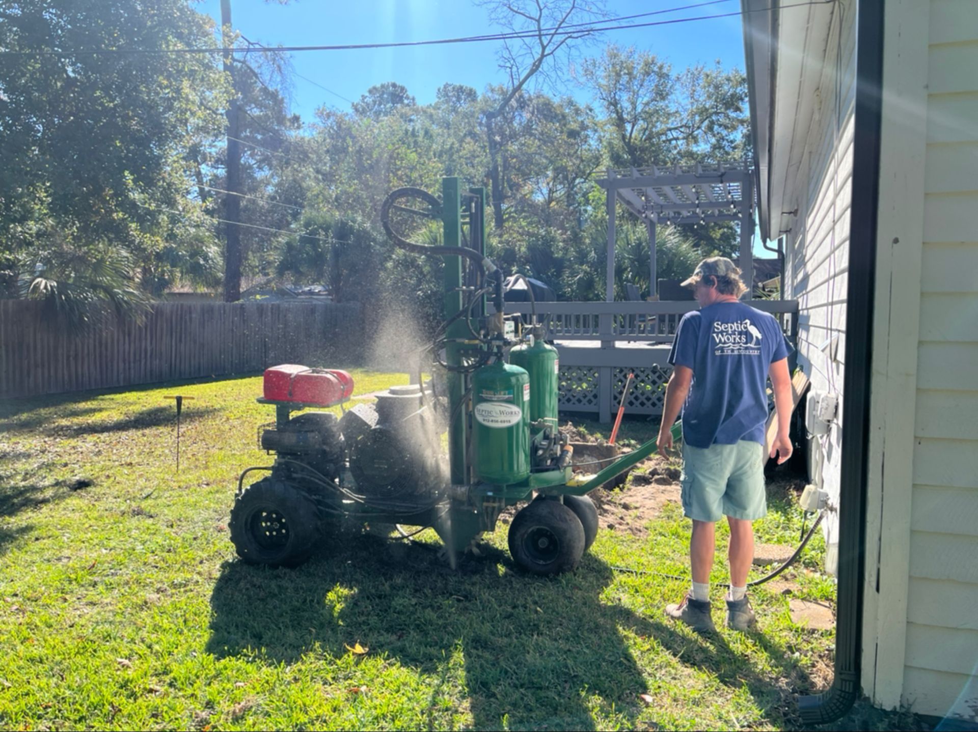 A man operates a green well-drilling machine in a grassy backyard, near a house with a gray deck.