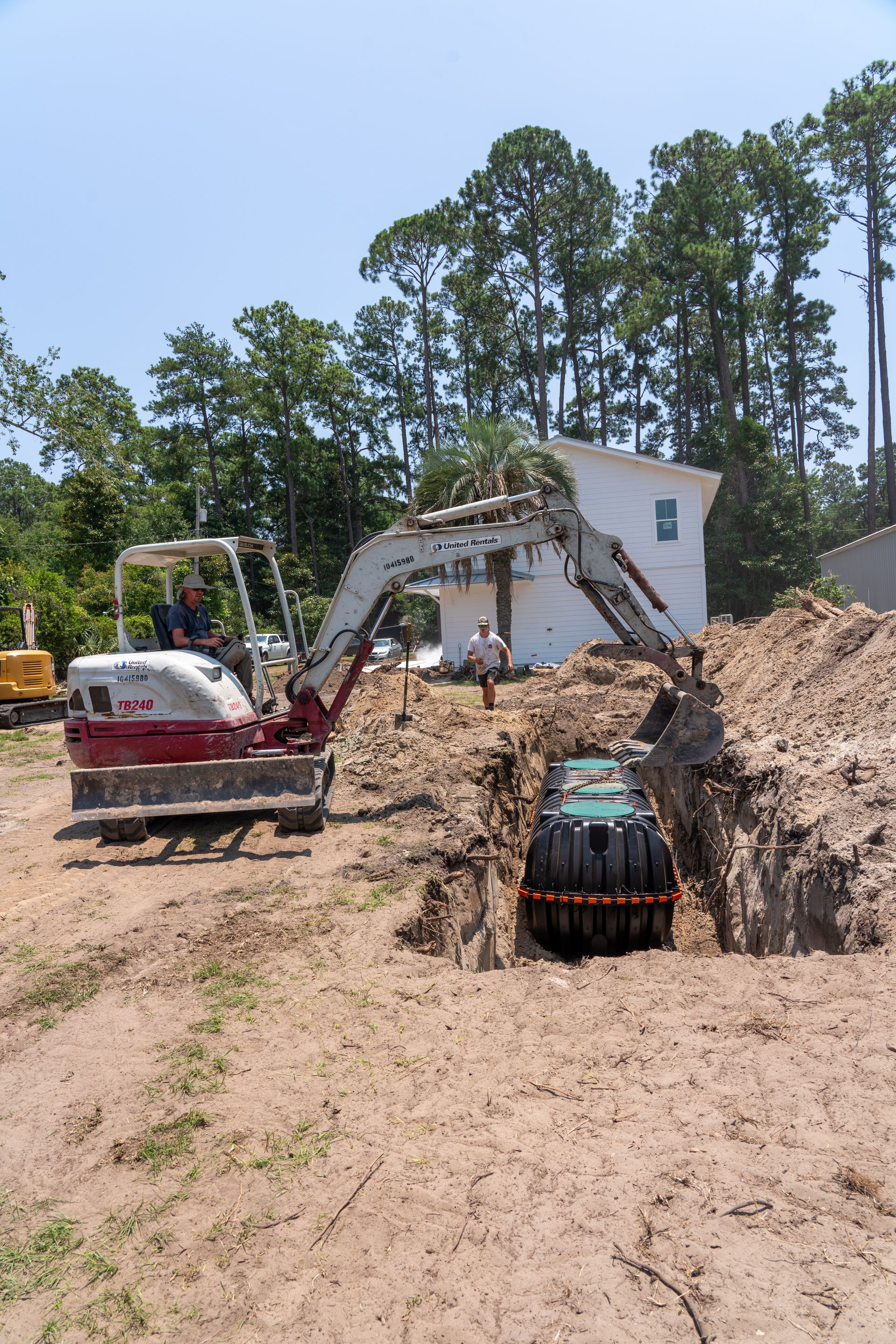 An excavator digs in a dirt pit, placing a large black tank. A building is in the background.