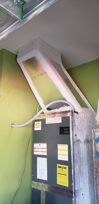 Electrical panel beneath white ducting in a green room, with exposed wiring and insulation damage.