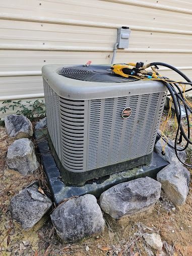 Outdoor air conditioning unit beside a house, surrounded by rocks, with tools and hoses on top