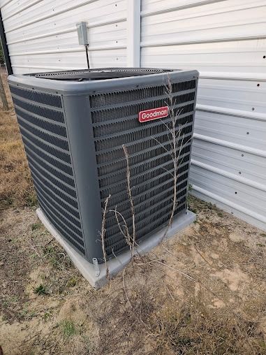Outdoor gray air conditioner unit beside a metal-sided building with dry grass and weeds nearby
