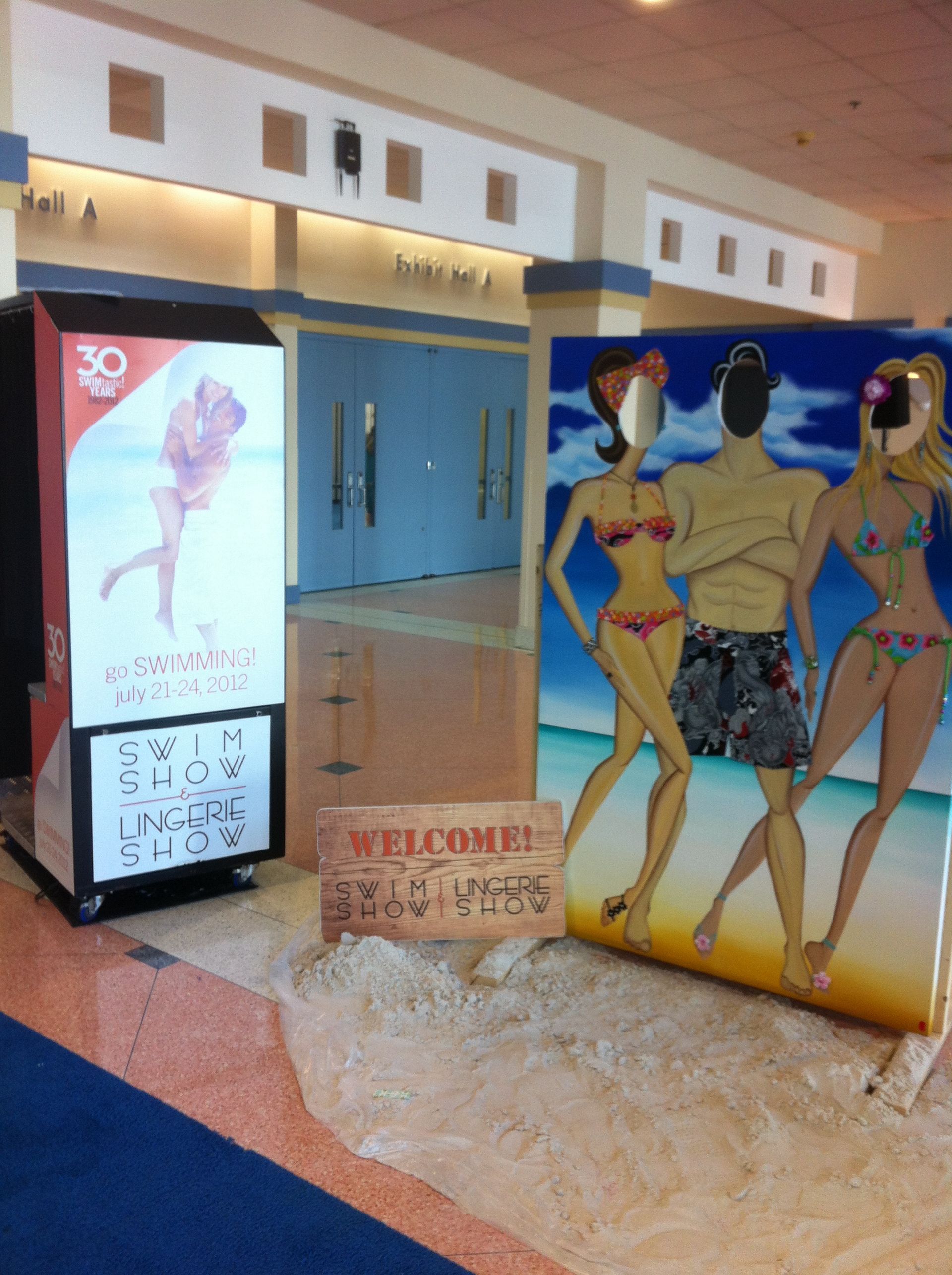 Beach-themed display in a building. Includes a photo op board and vendor kiosk on a sand-like surface.