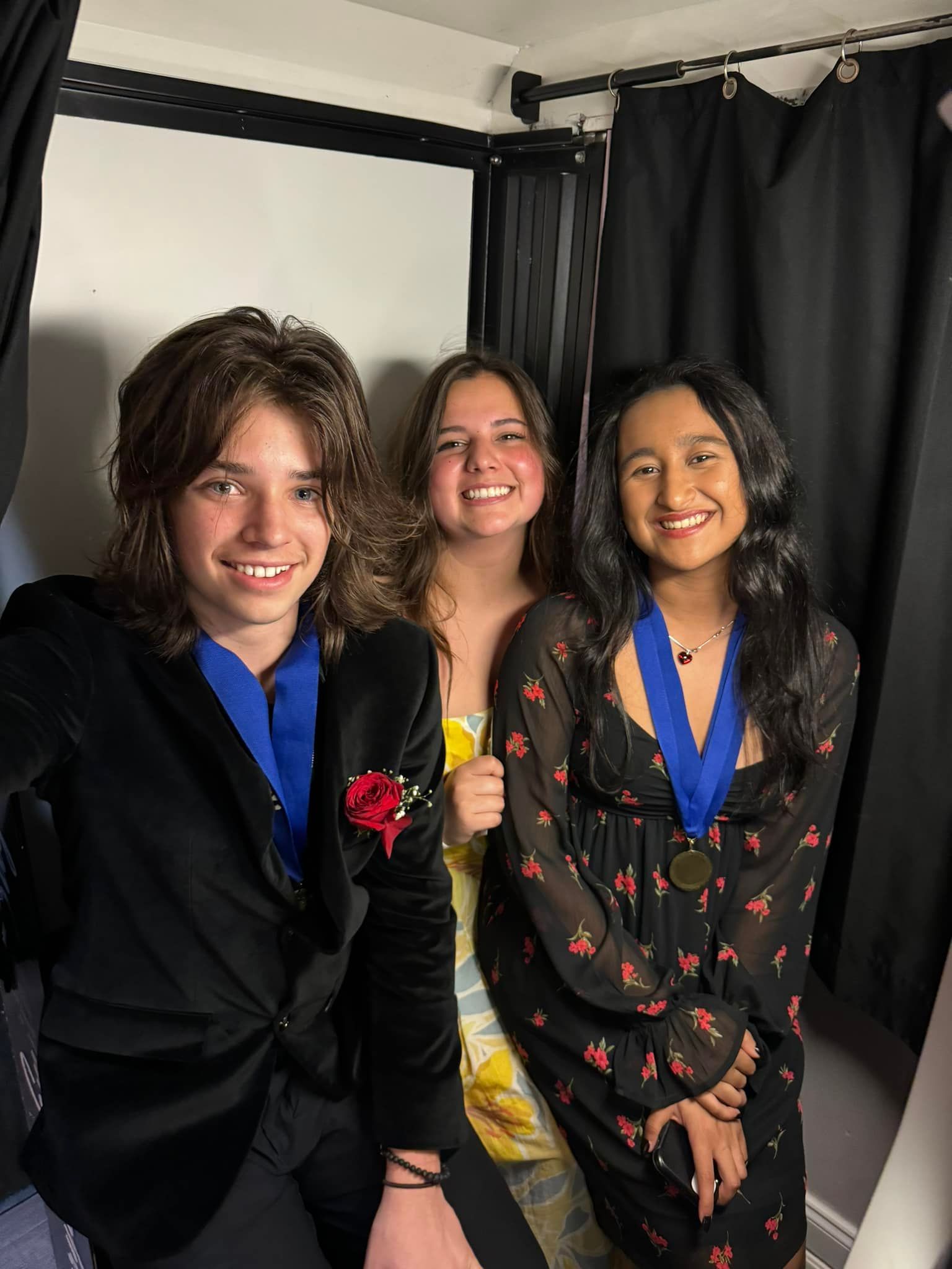 Three young people smiling, wearing formal attire and medals, posing near black curtains.