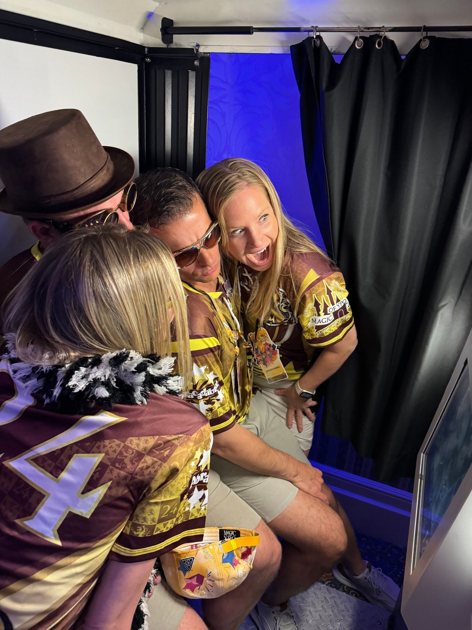 Four people posing in a photo booth, wearing gold and brown shirts, smiling, one with a top hat.