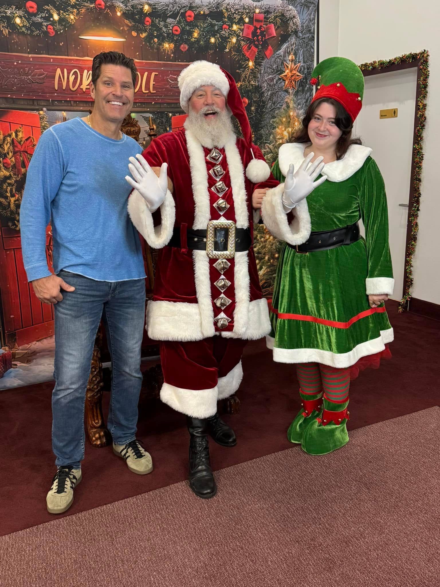 Man, Santa Claus, and elf pose in front of a Christmas backdrop. Santa waves with wide smile, elf gestures, man smiles.