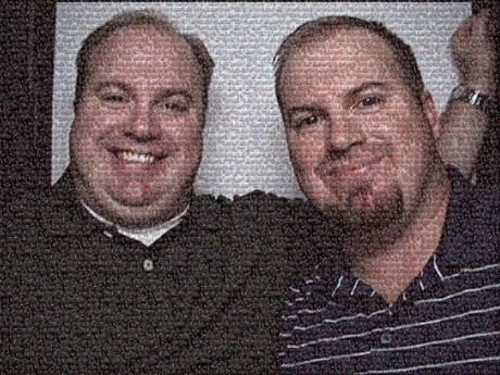 Two men smiling for a photo. One has a full beard, the other a clean-shaven face, both in a dark setting.