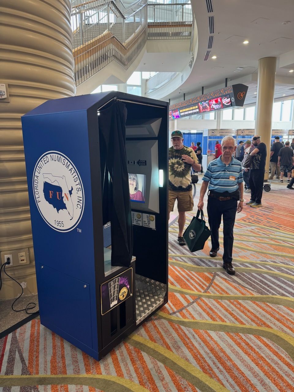 Photo booth in a convention center hallway; two men walk by.