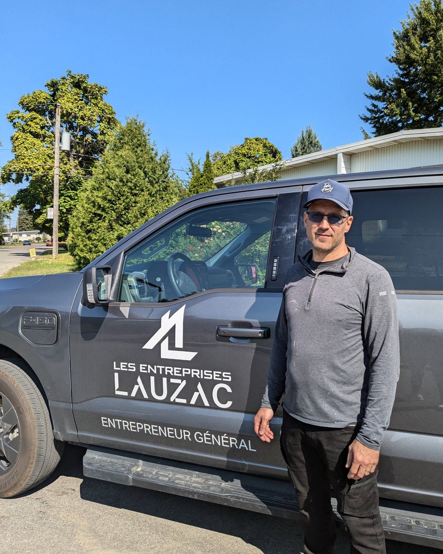 Un homme portant une casquette et une chemise grise se tient à côté d'un camion portant le logo de l'entreprise « Les Entreprises Lauzac ».