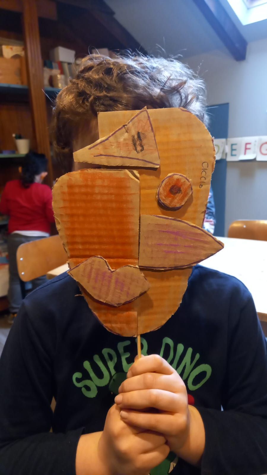 Un ragazzo che indossa una maglietta da super dinosauro tiene una maschera di cartone davanti al viso