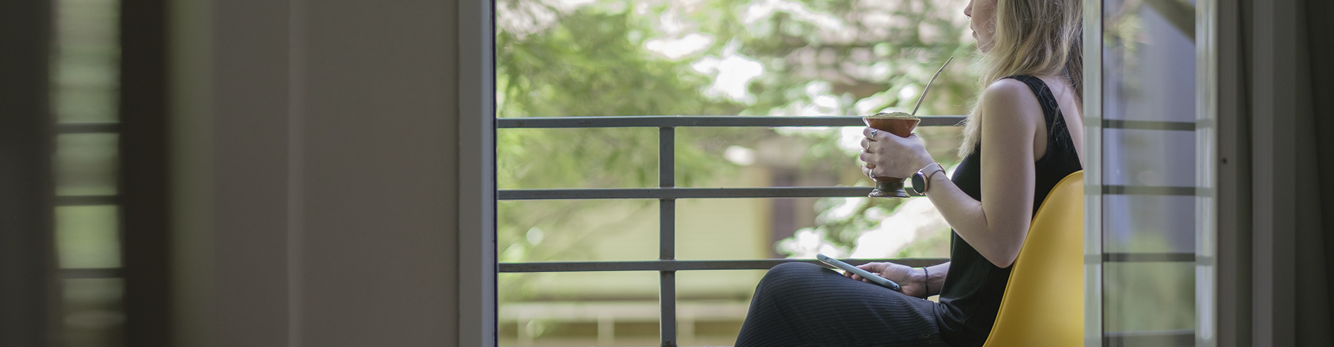 Uma mulher está sentada em uma varanda bebendo vinho e olhando pela janela.