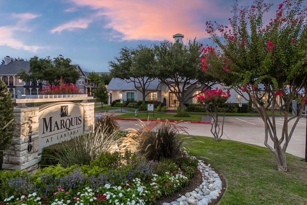 a sign for the marquis apartment complex is surrounded by flowers and trees at Marquis at Lantana in Flower Mound, TX.