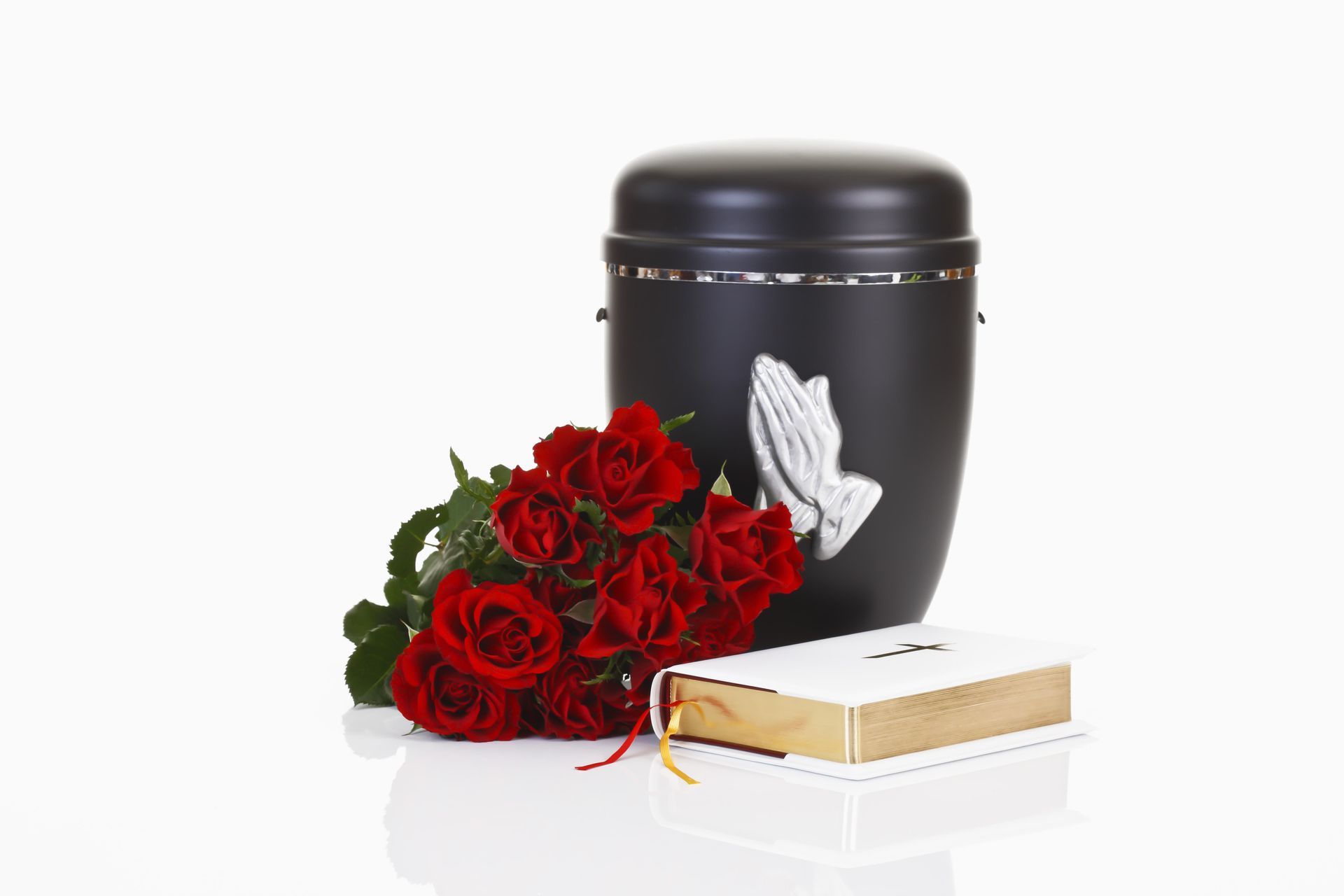 Black urn with praying hands, red roses, and a Bible on a white surface.
