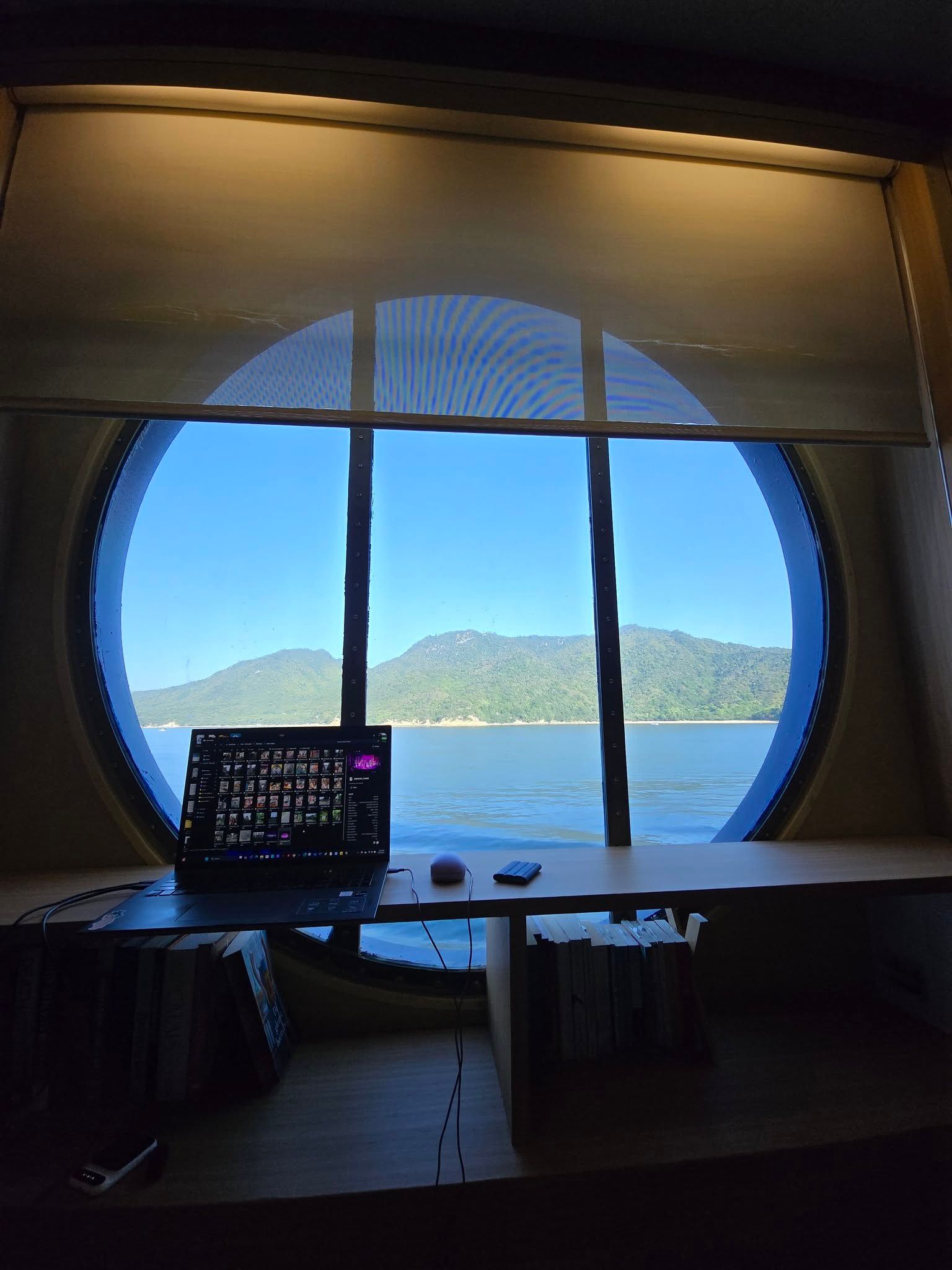 A laptop sits on a desk in front of a round window overlooking a body of water