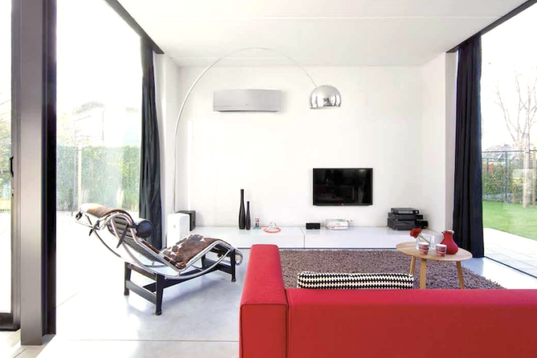 A residential HVAC is installed on the top of a white wall of the living room near the tv. The other sides of the walls are replaced by full-length windows