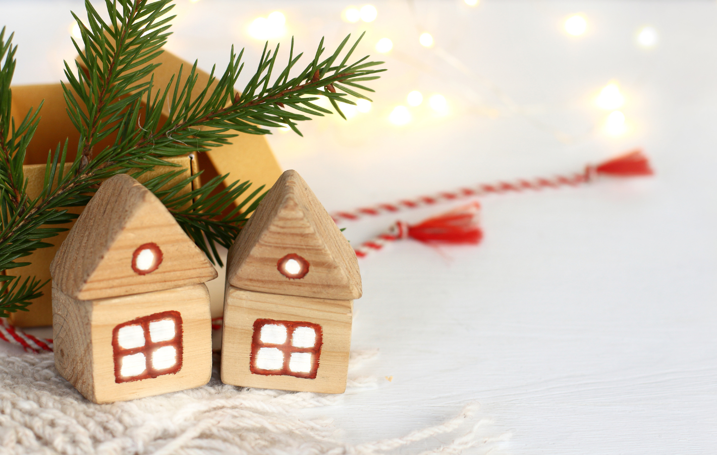 Two wooden houses against the background of a spruce branch, an opened gift box, and garland lights.