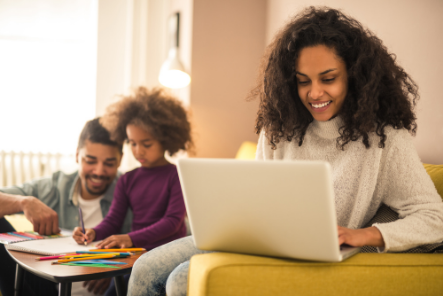 Picture of a woman working from her laptop with her husband and daughter playing next to her.