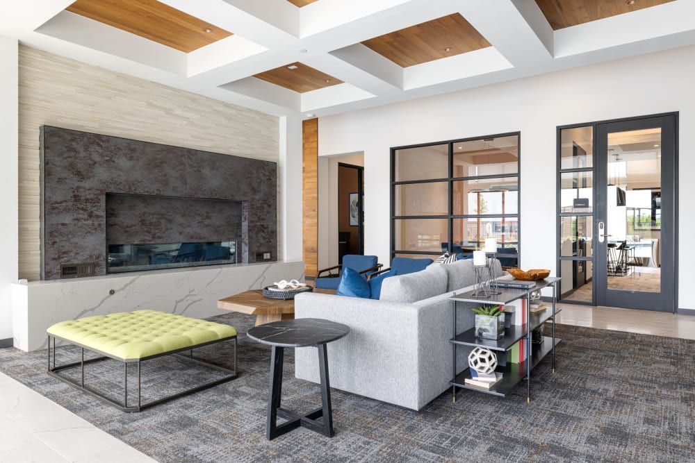 Modern clubhouse interior featuring a gray sectional sofa with blue pillows, a wall-mounted TV within a dark textured wall, and a horizontal fireplace below. An olive green tufted ottoman, a dark round side table, and a glass coffee table with decorative items and books rest on a patterned area rug. Exposed wooden beams accent the high ceiling, and natural light floods through the glass doors leading to an adjoining room with a table set visible at Marq Perimeter in Atlanta, GA.