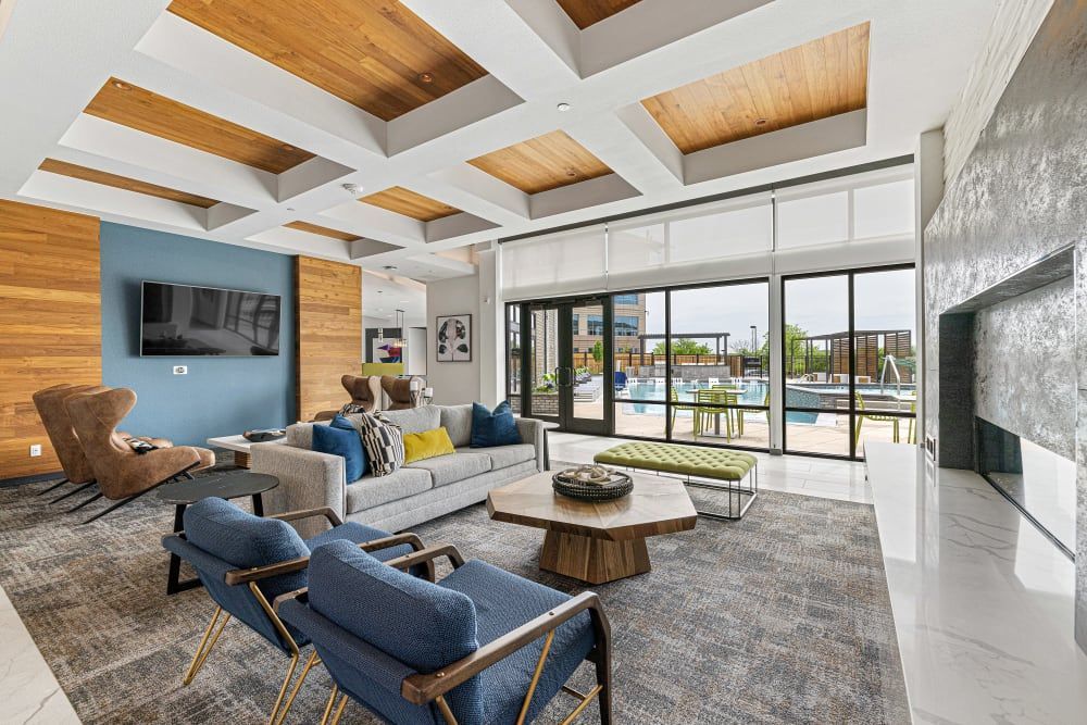 Modern clubhouse with coffered ceiling featuring exposed wooden beams, an accent wall painted in blue with a mounted flat-screen TV, and wood-panel wall. Contemporary furniture including a grey sectional sofa with colorful pillows, two blue armchairs, a brown suede chair, and a central wooden coffee table. Large glass sliding doors provide a view to an outdoor pool area. Fireplace with a white marble-like surround. Decorated with subtle art pieces and plush textures, reflecting a stylish interior design at Marq Perimeter in Atlanta, GA.