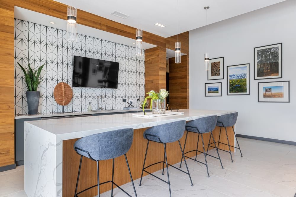 Modern clubhouse kitchen with a geometric tile backsplash and wooden accents. The breakfast bar features a white marble countertop with four blue upholstered bar stools with black metal legs. Above the bar hang elegant glass pendant lights. A wall-mounted flat-screen TV is centered between two green potted plants. To the right, framed landscape photographs decorate the light gray wall at Marq Perimeter in Atlanta, GA.