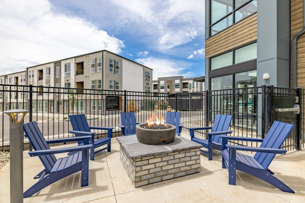 Outdoor communal area with a lit fire pit surrounded by blue Adirondack chairs, with modern apartment buildings in the background and a partly cloudy sky above at Marq Perimeter in Atlanta, GA.