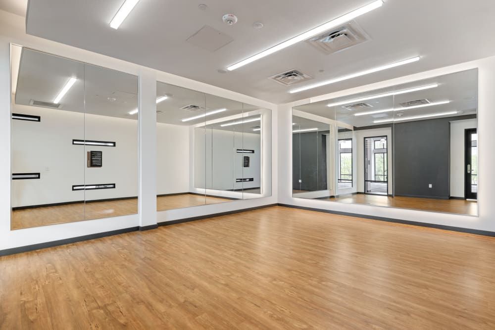 A spacious and well-lit dance studio with large mirrors lining one wall, wooden floors, and a barre along the mirrored wall. The space is empty and features a white ceiling with fluorescent lights and grey painted walls with back exit doors at Marq Perimeter in Atlanta, GA.