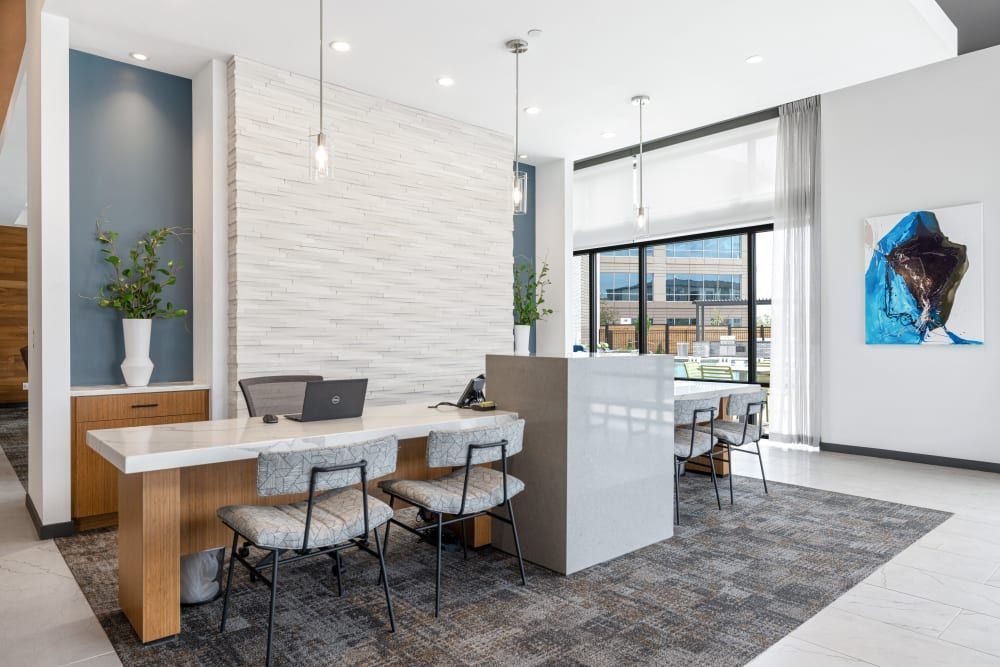 Modern leasing office with clean lines and contemporary design. Features a front desk with a laptop, marble accents, and comfortable seating areas. White textured wall, blue accents, large windows with sheer curtains, and an abstract blue artwork on the wall add to the stylish interior at Marq Perimeter in Atlanta, GA.