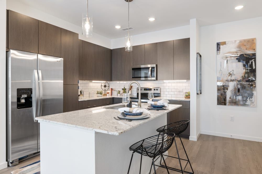 Modern kitchen interior with stainless steel appliances, including a side-by-side refrigerator, microwave, and oven range hood. The kitchen features dark wood cabinets, white subway tile backsplash, and a central island with a marble countertop and two black bar stools. Pendant lights hang above the island, and an abstract painting decorates the wall at Marq Perimeter in Atlanta, GA.