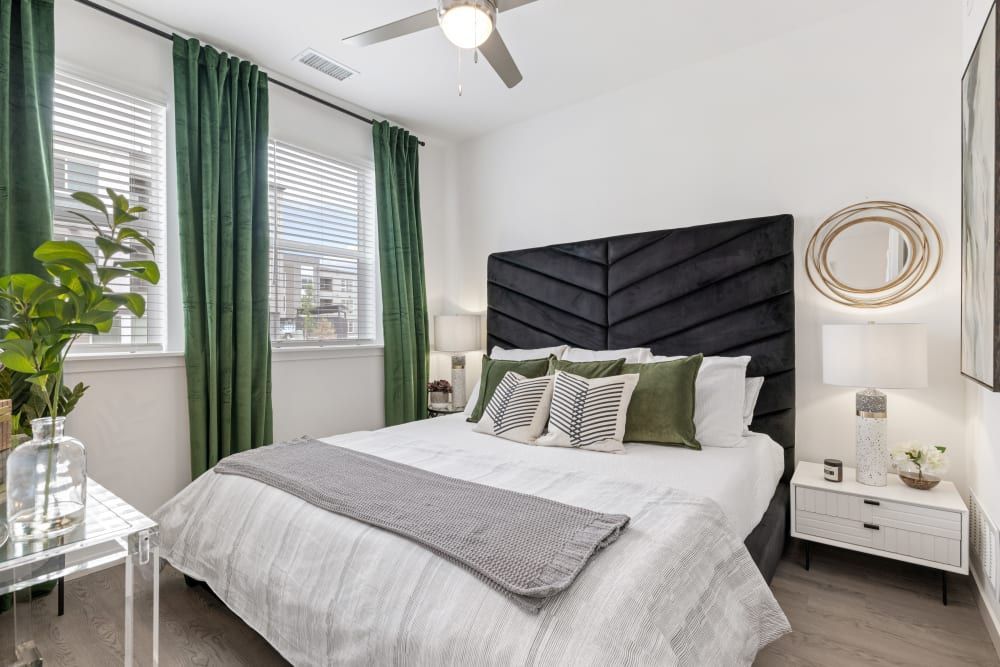 A modern and stylish bedroom featuring a large bed with a black tufted headboard against a white wall. The bed is dressed in white linen with a gray throw blanket and decorative cushions in shades of green and patterned black and white. To the right, a round gold mirror hangs above a white nightstand with a contemporary lamp and a small plant. The room is well-lit by natural light streaming in from two windows dressed with floor-length emerald green curtains. A ceiling fan is present above the bed, and to the left, there is a clear acrylic side table with a potted plant, contributing to a crisp and clean ambiance. The flooring is light wood, complementing the neutral and green color scheme at Marq Perimeter in Atlanta, GA.