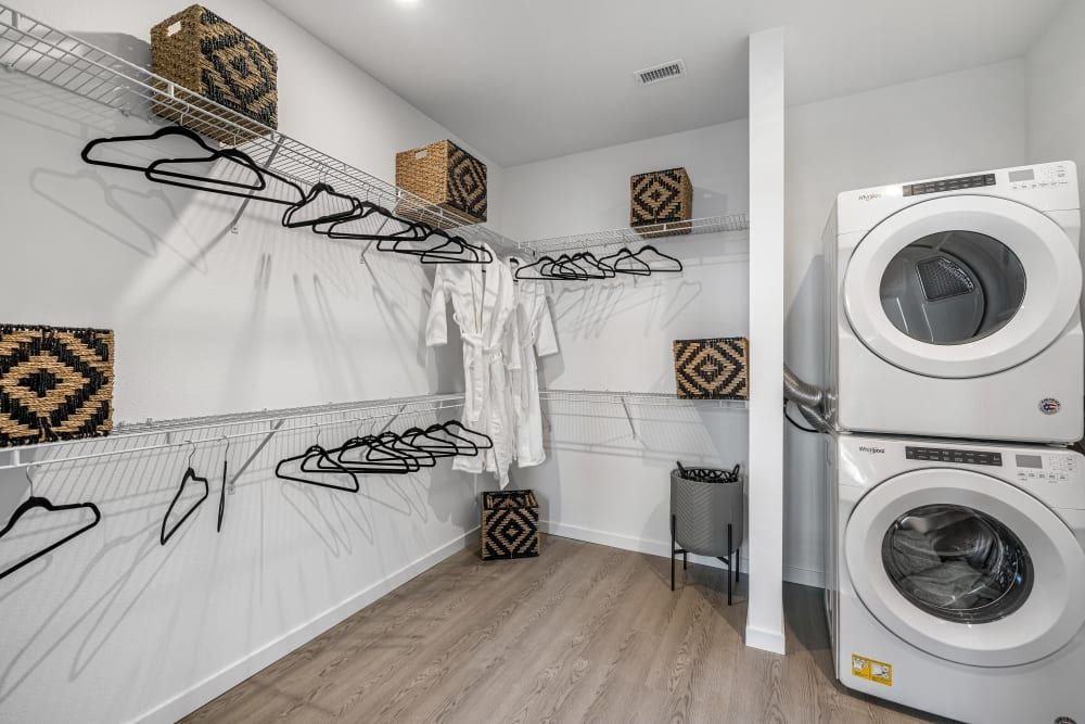 A modern laundry room with white walls and wooden flooring featuring a stacked tumble dryer over a washing machine on the right. Wire shelving runs along the left wall with empty hangers and a few wicker baskets, and a single white bathrobe hangs amongst the hangers. A black laundry basket sits on the floor to the right of the machines at Marq Perimeter in Atlanta, GA.