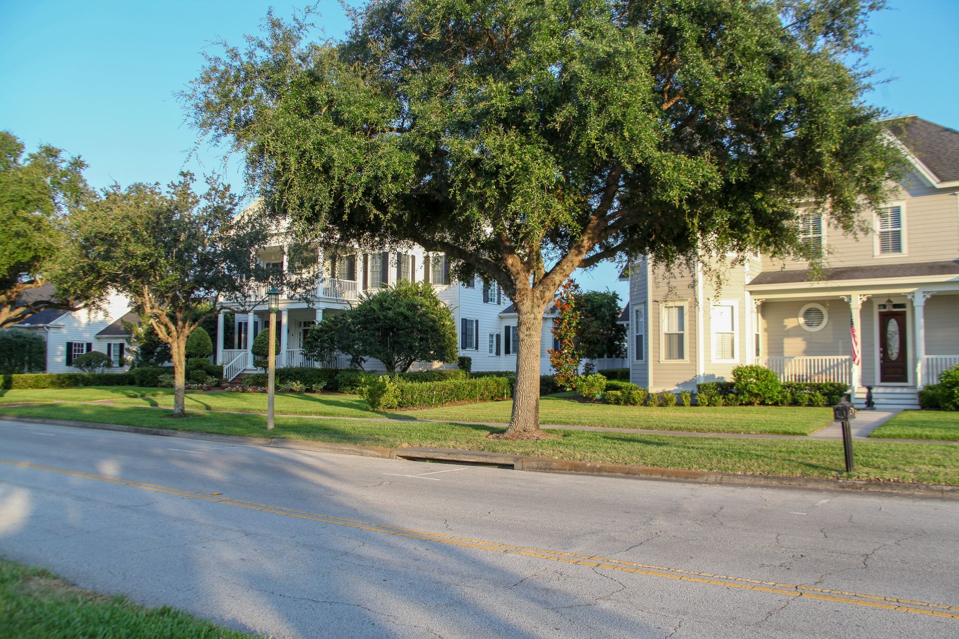 Two large houses with porches sit on a sunny, green residential street under a clear blue sky.