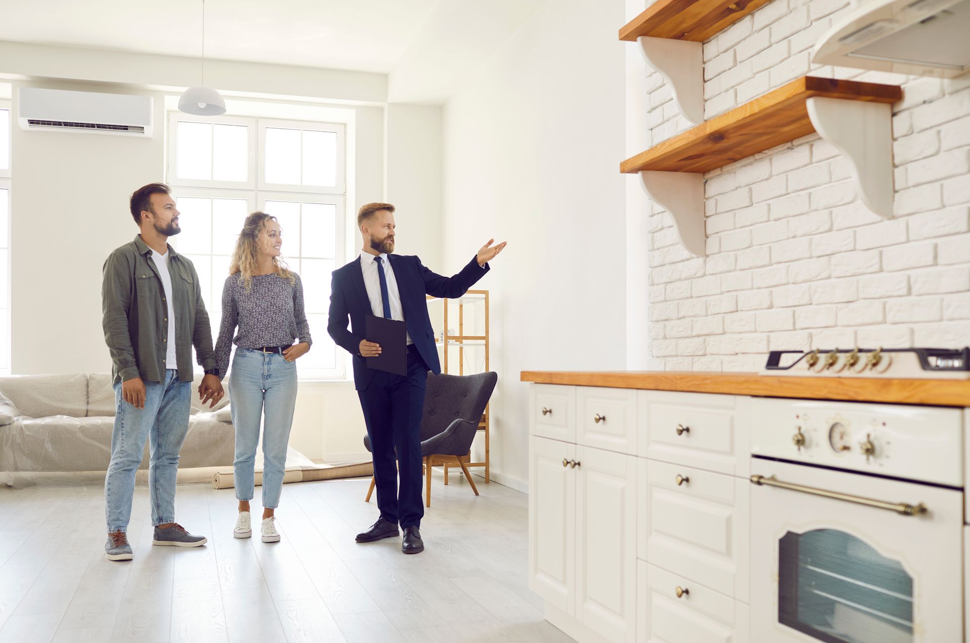 A real estate agent gestures toward a kitchen wall while showing a home to a couple.