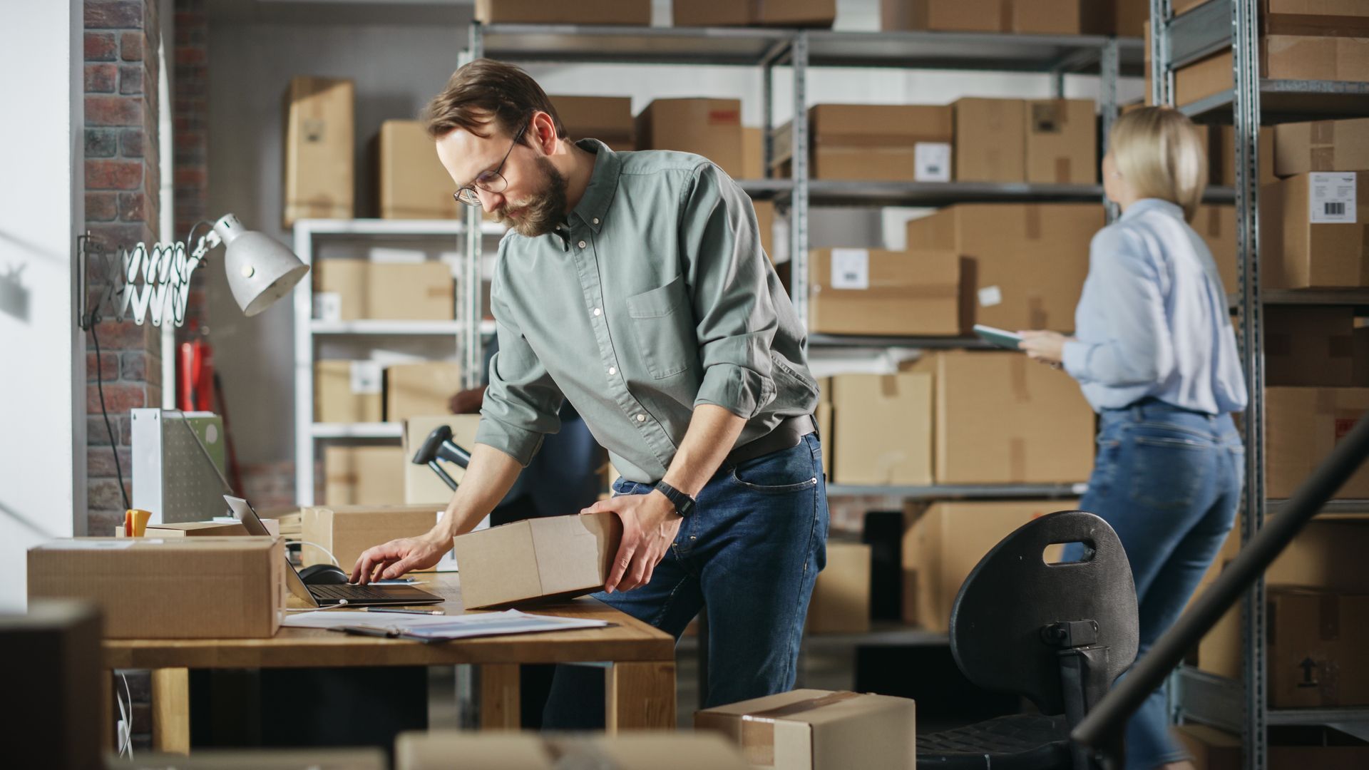 A person focuses on packing a cardboard box on a desk in a warehouse, while another person stands by nearby shelves.