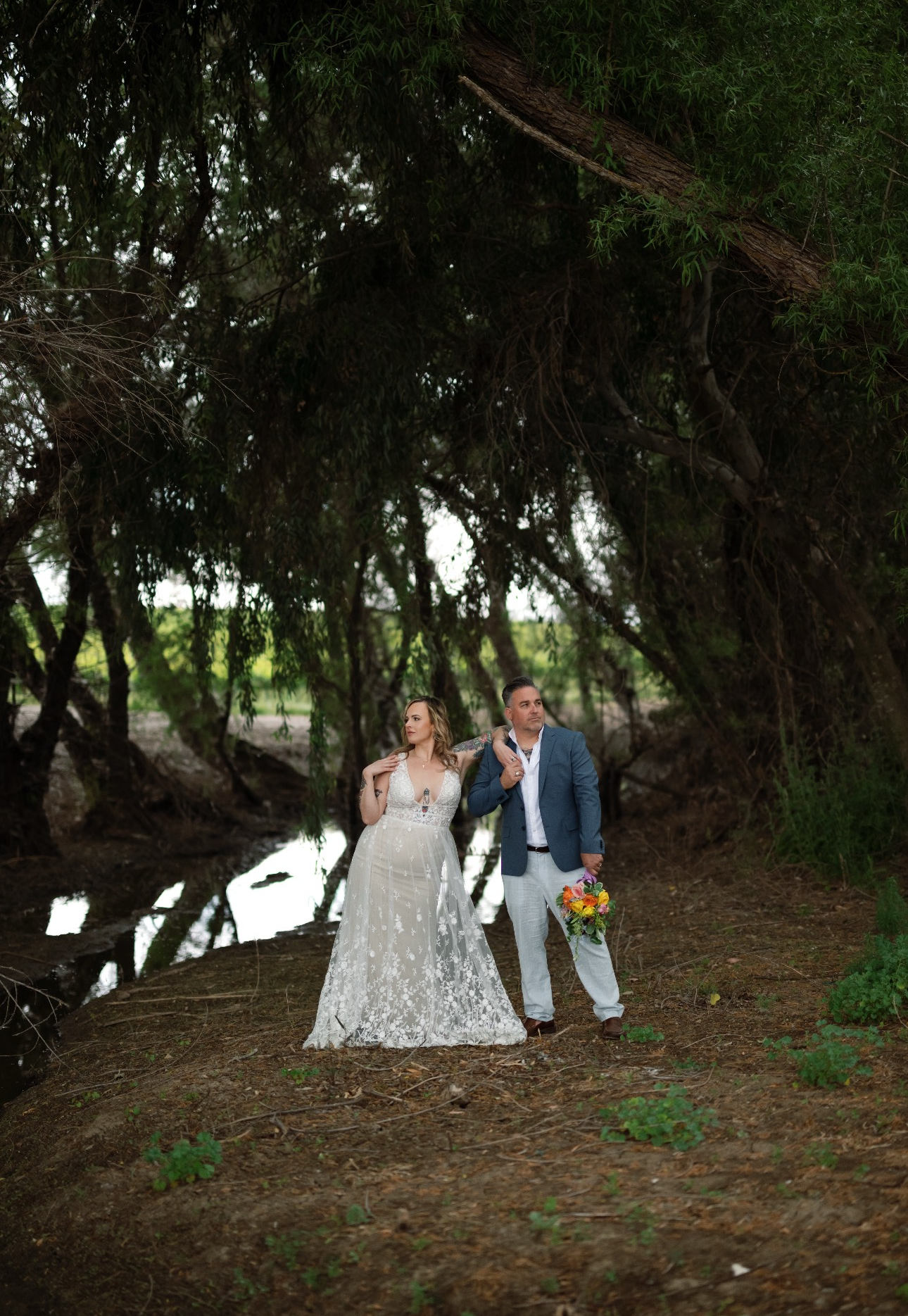 Bride and groom stand near water under a canopy of trees. The woman wears a white wedding gown; the man wears a blue blazer.