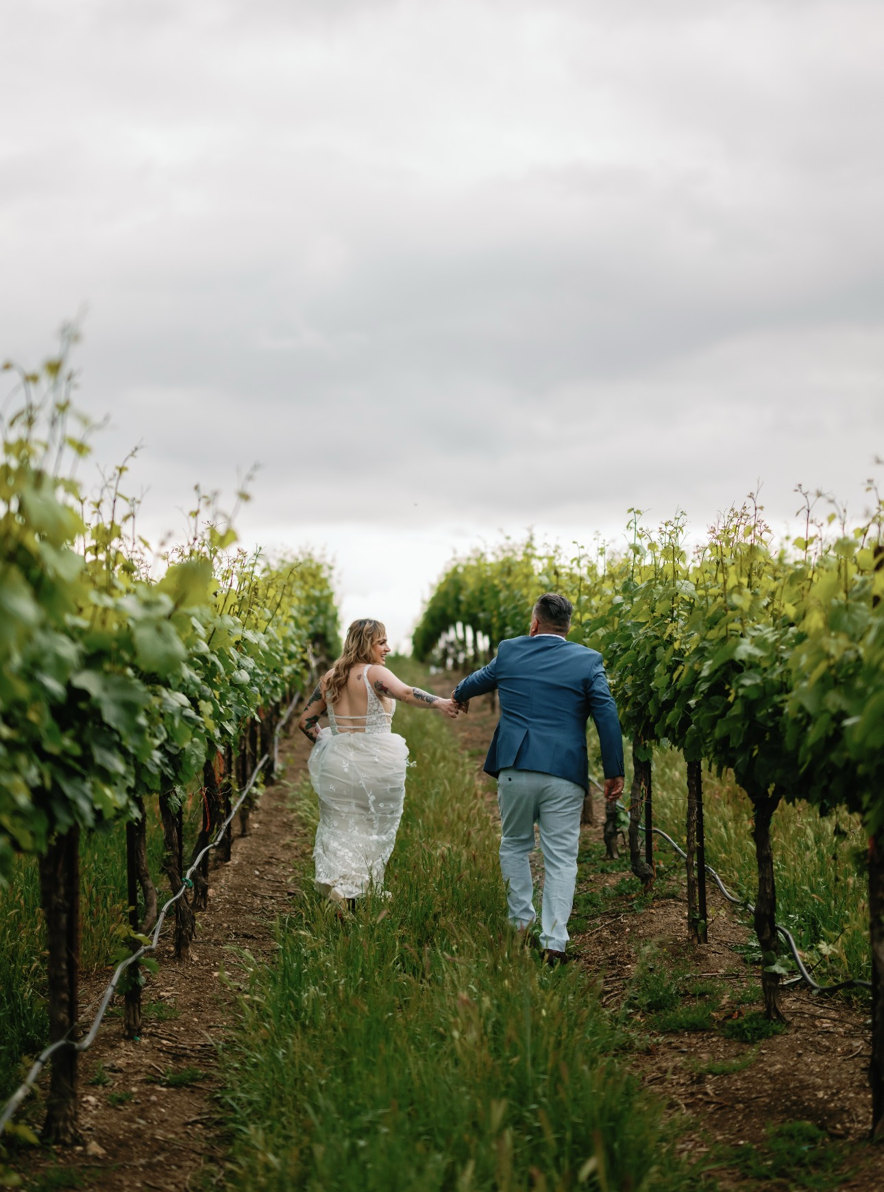 A newly married couple walks hand-in-hand through a vineyard, bride in white, groom in blue jacket, under a cloudy sky.