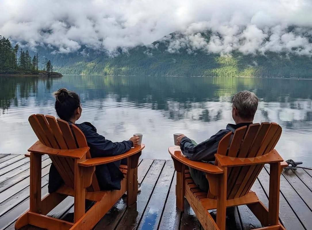 Twee mensen zitten op houten stoelen op een steiger en kijken uit over een mistig meer en beboste bergen.