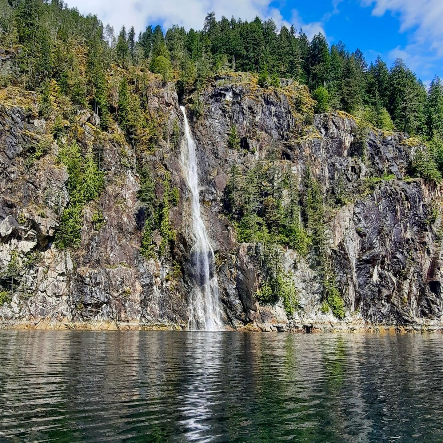Een waterval stort zich van een rotsachtige klif naar beneden in een kalm meer onder een blauwe hemel met bomen op de top.