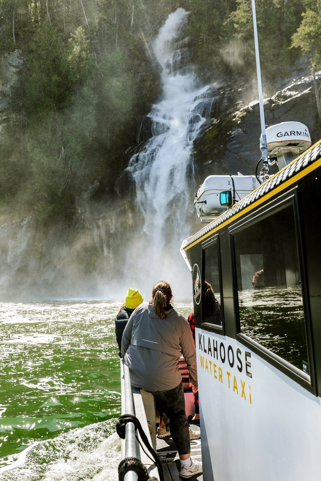 Mensen in een boot bij een waterval, met opstijgende mist en een ander vaartuig op de achtergrond.