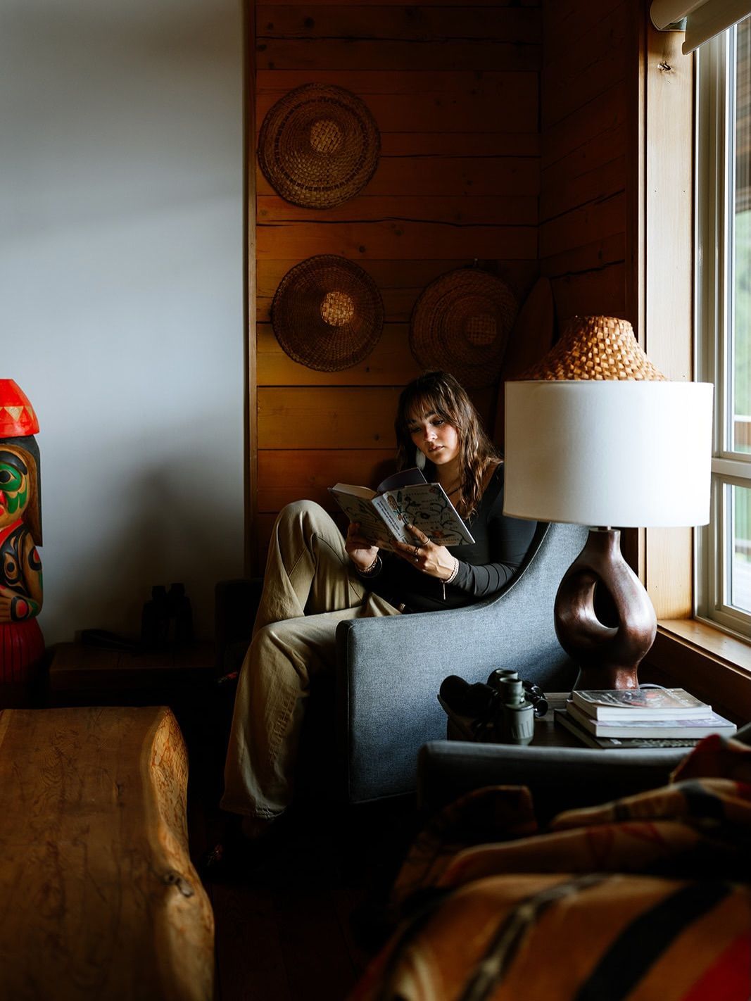 Iemand zit in een gezellige woonkamer bij het raam, te lezen op een bank naast een lamp.