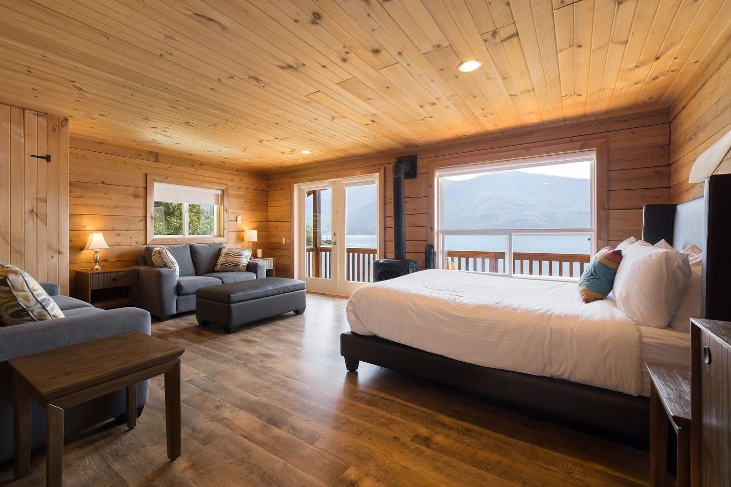 Gezellige houten slaapkamer met een bed, een bank en grote ramen met uitzicht op het water.