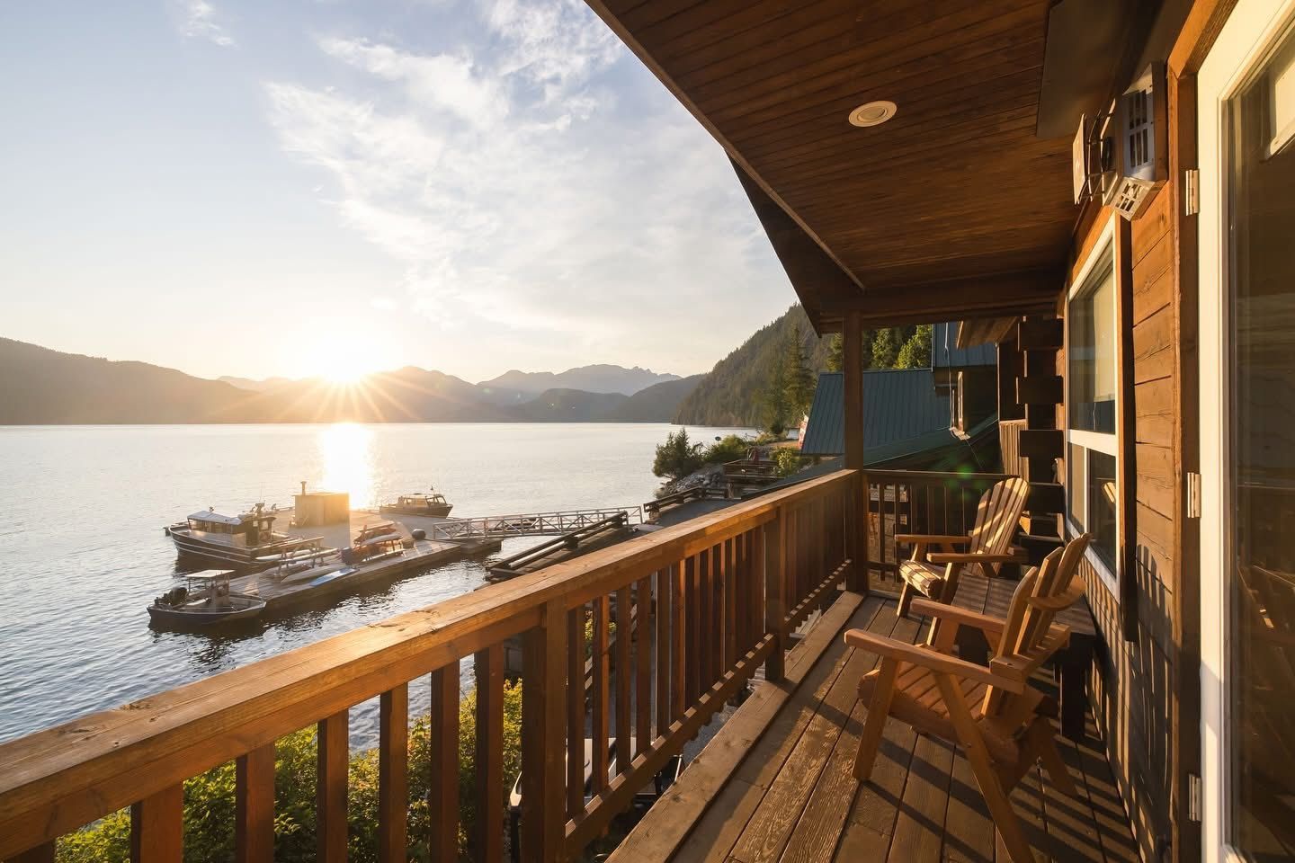 Zonsopgang boven een meer, gezien vanaf het balkon van een houten blokhut met stoelen en aangemeerde boten.