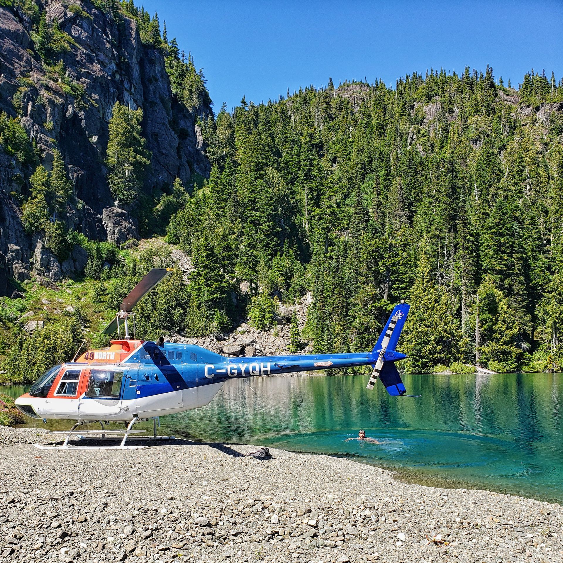 Een blauw-witte helikopter staat geparkeerd op een rotsachtige rivieroever naast helder turkooizen water en beboste kliffen.