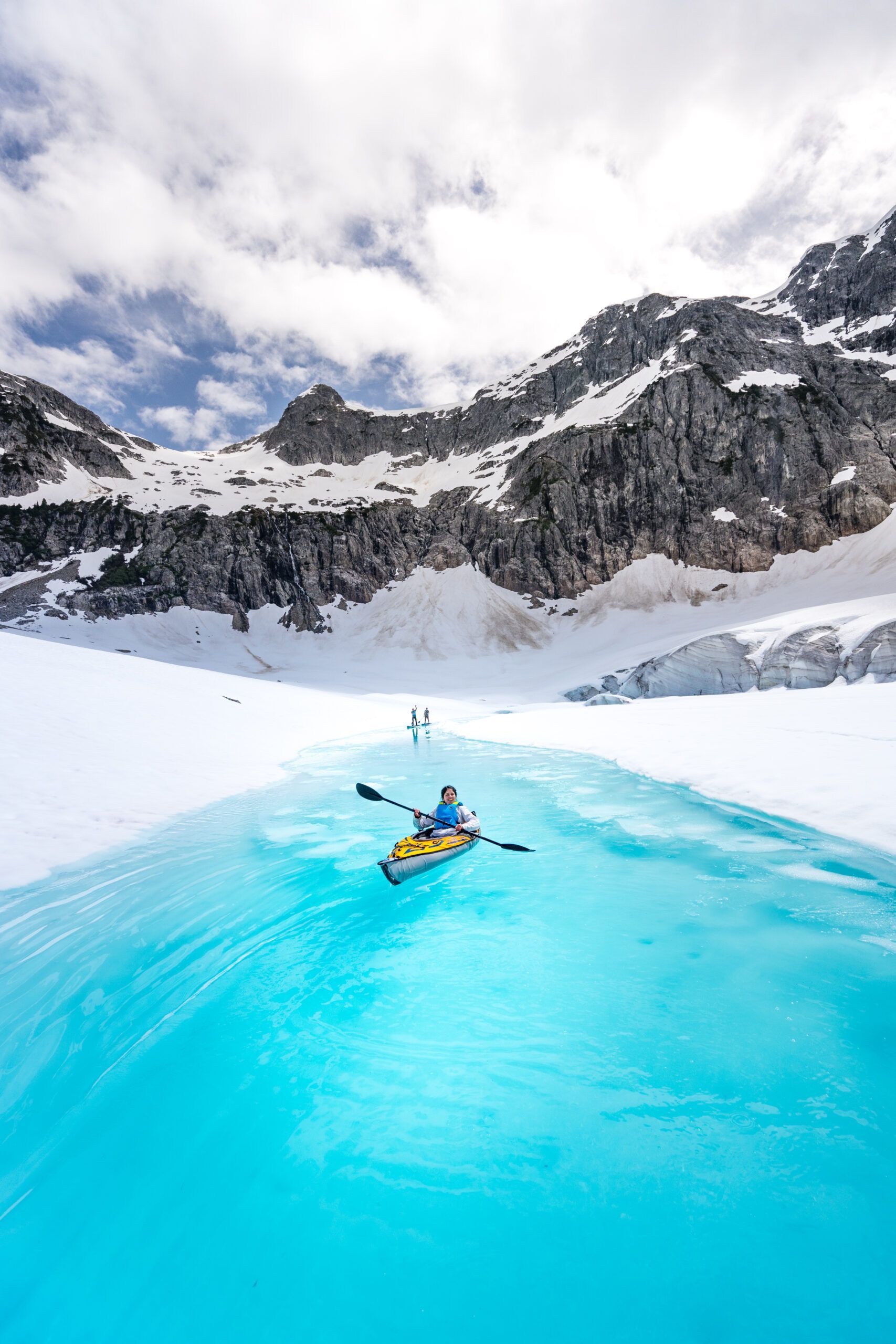 Een kajakker vaart op helder turkooizen gletsjerwater onder besneeuwde bergen en een bewolkte hemel.