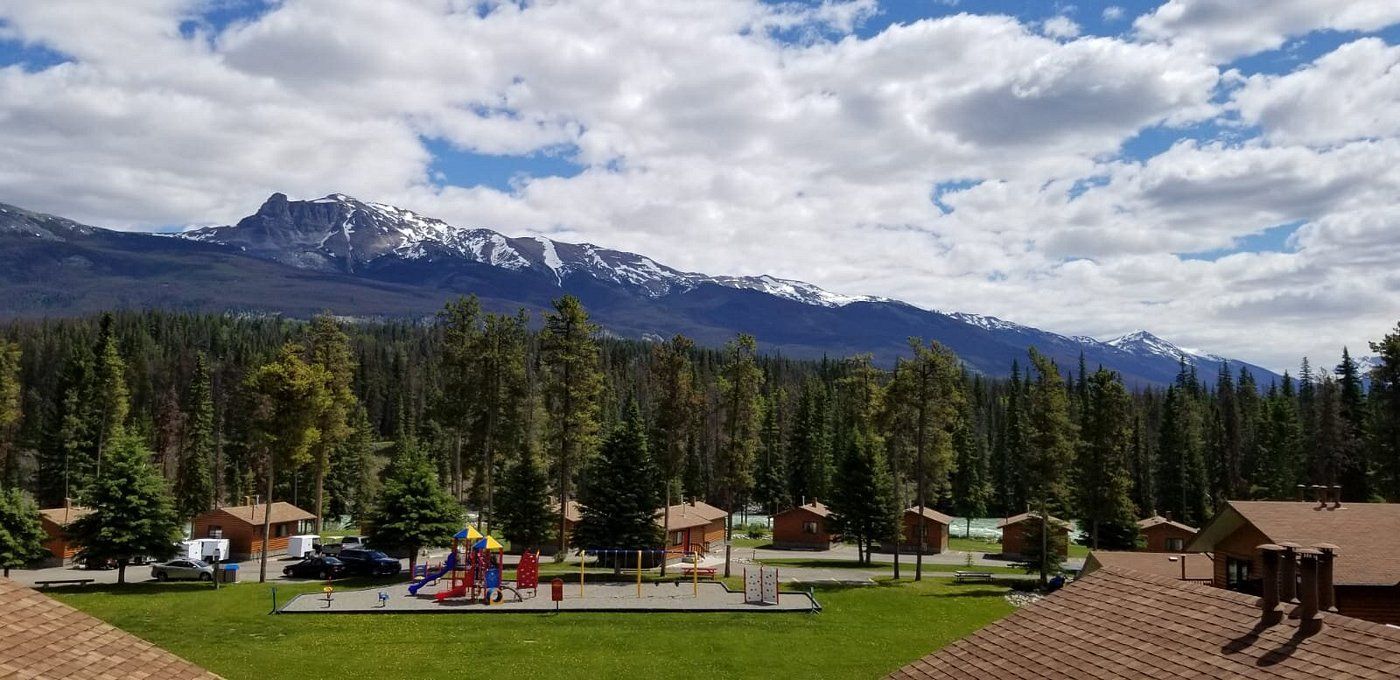 Overzicht van Becker's Chalets Jasper met een speeltuin, dennenbomen en besneeuwde bergtoppen onder een bewolkte hemel.