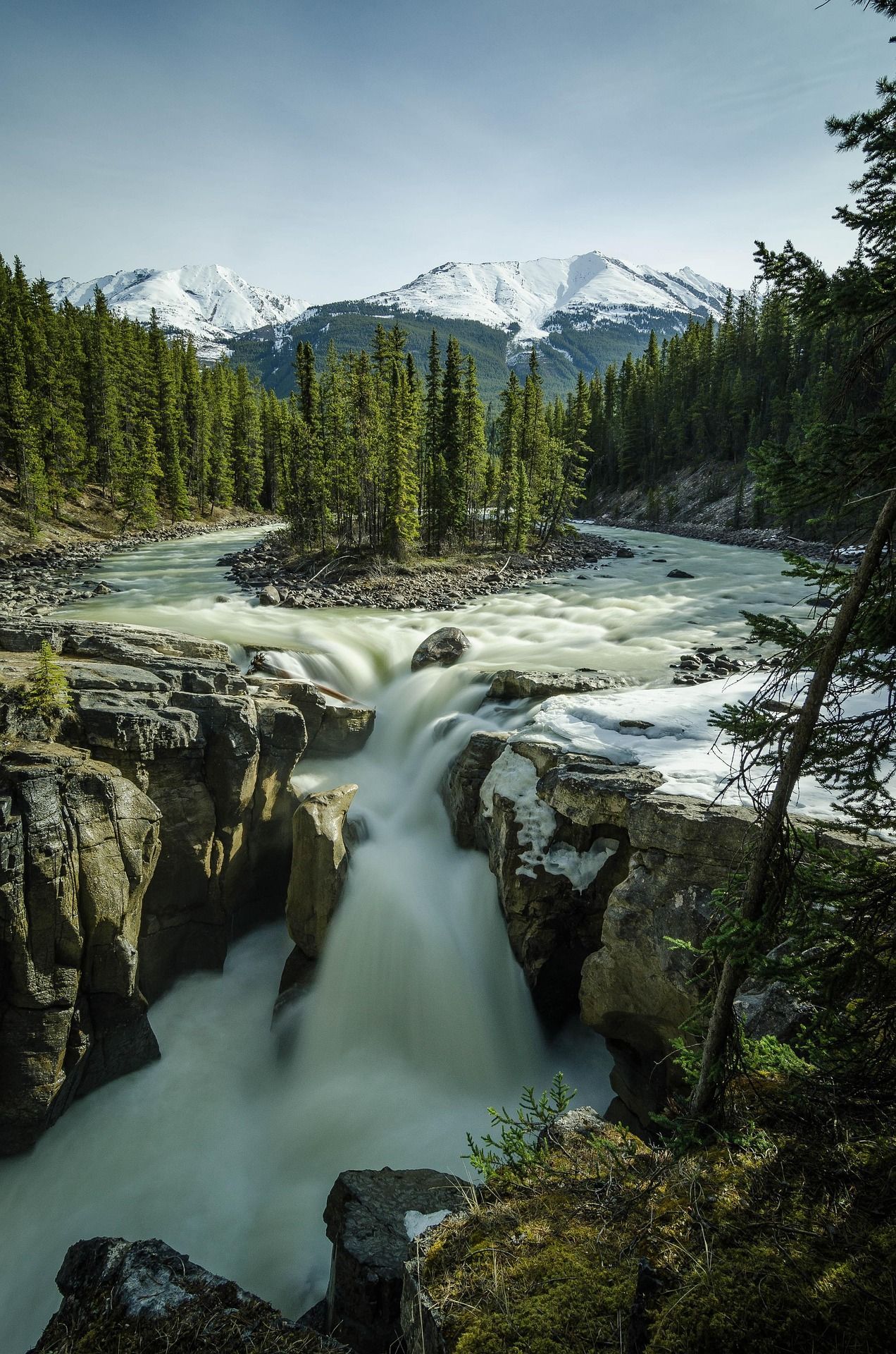 Waterfall cascading over rocks with snow-capped mountains and evergreen trees in the background.