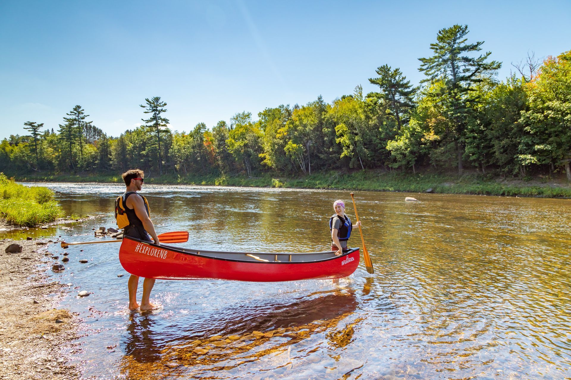 Two people with a red canoe in a shallow river, surrounded by trees under a bright sky.