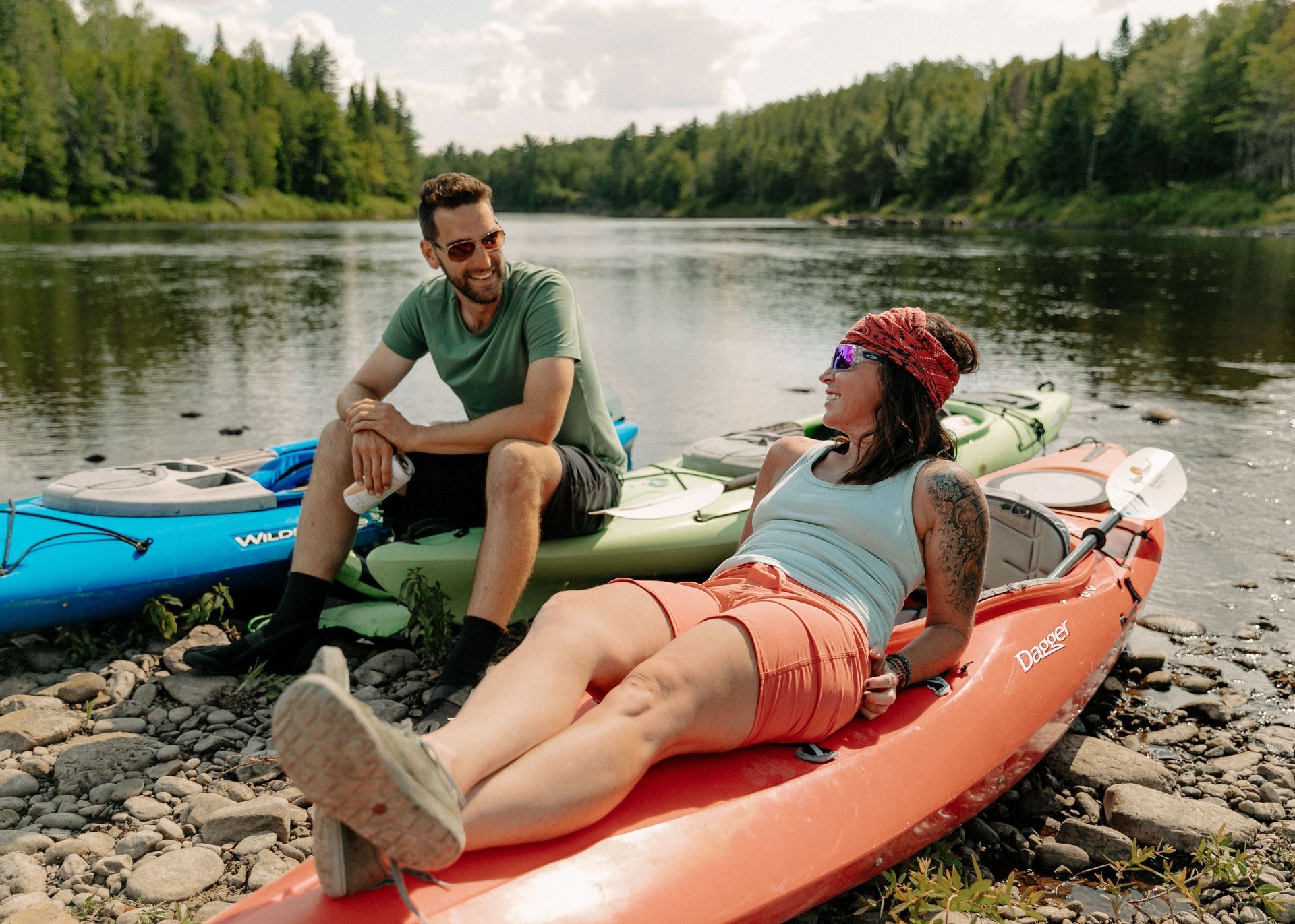 Two people relaxing on kayaks by a river, one drinks from a can, sunny day, green forest backdrop.