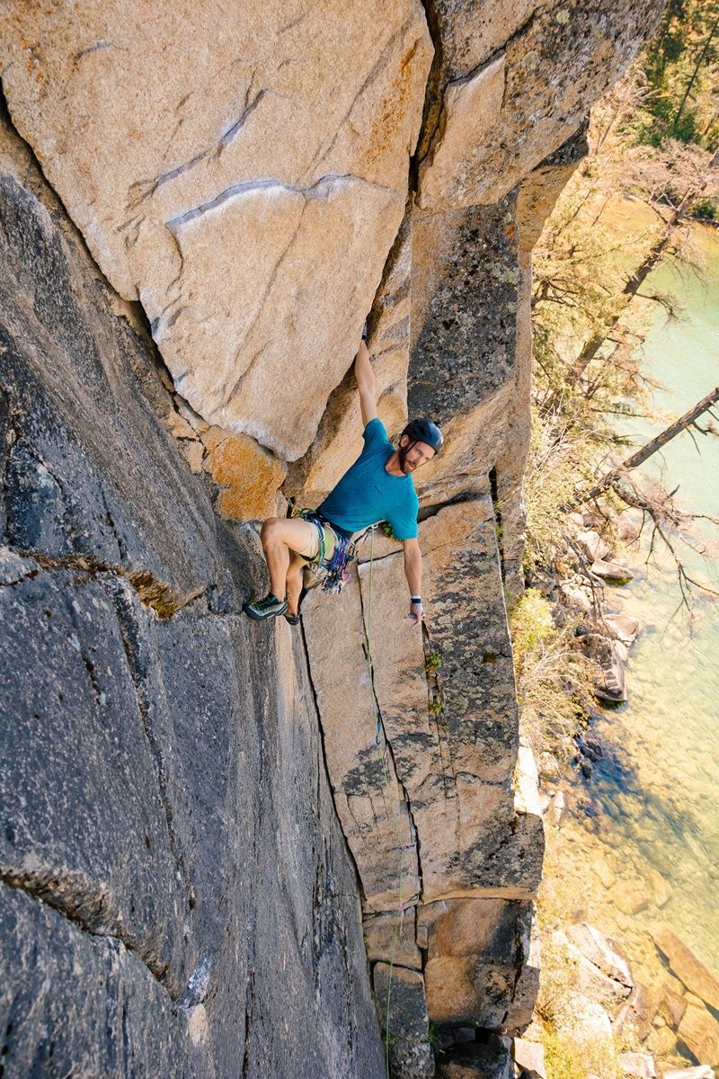 Rock climber ascending a steep cliff face; blue shirt, light skin, turquoise water below.