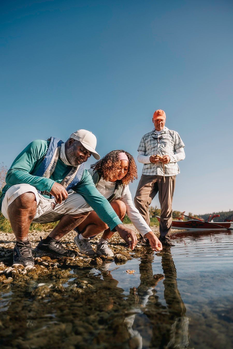 Drie mensen onderzoeken ondiep water. Eén wijst, de anderen kijken toe. Buiten, zonnig weer.