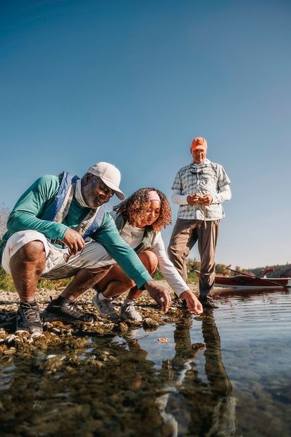 Drie mensen onderzoeken ondiep water. Eén wijst, de anderen kijken toe. Buiten, zonnig weer.