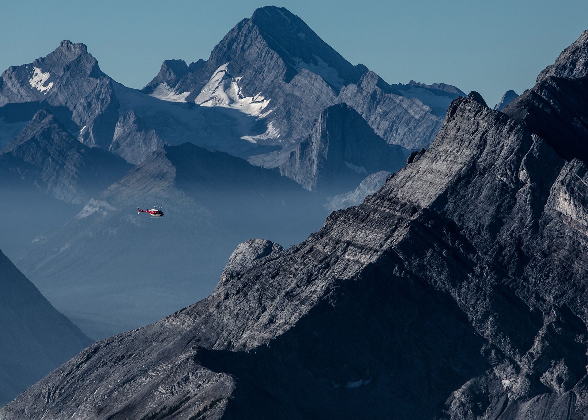 Ruige bergtoppen met een kleine rode helikopter in de lucht tegen een blauwe hemel.